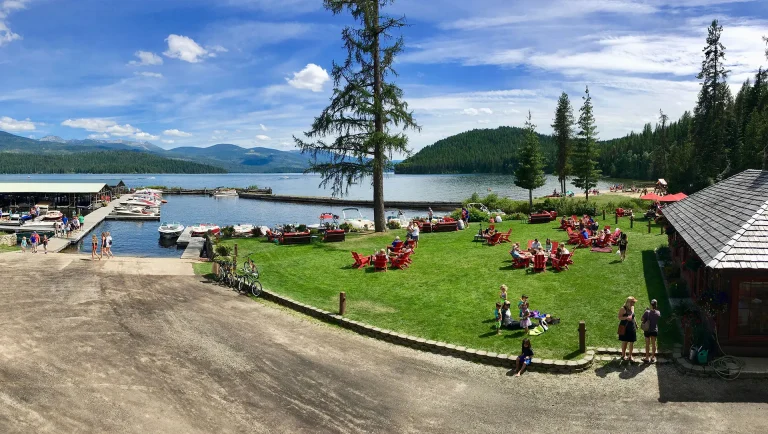 people gathered by the lake on a sunny day sitting on the grass in red chairs by boats at the dock at Elkins Resort in Priest Lake